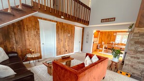 Un vaste espace de vie comprenant un canapé rouge, un divan en cuir noir et une table basse en bois. Un escalier mène à un niveau supérieur, et on aperçoit une cuisine au fond.