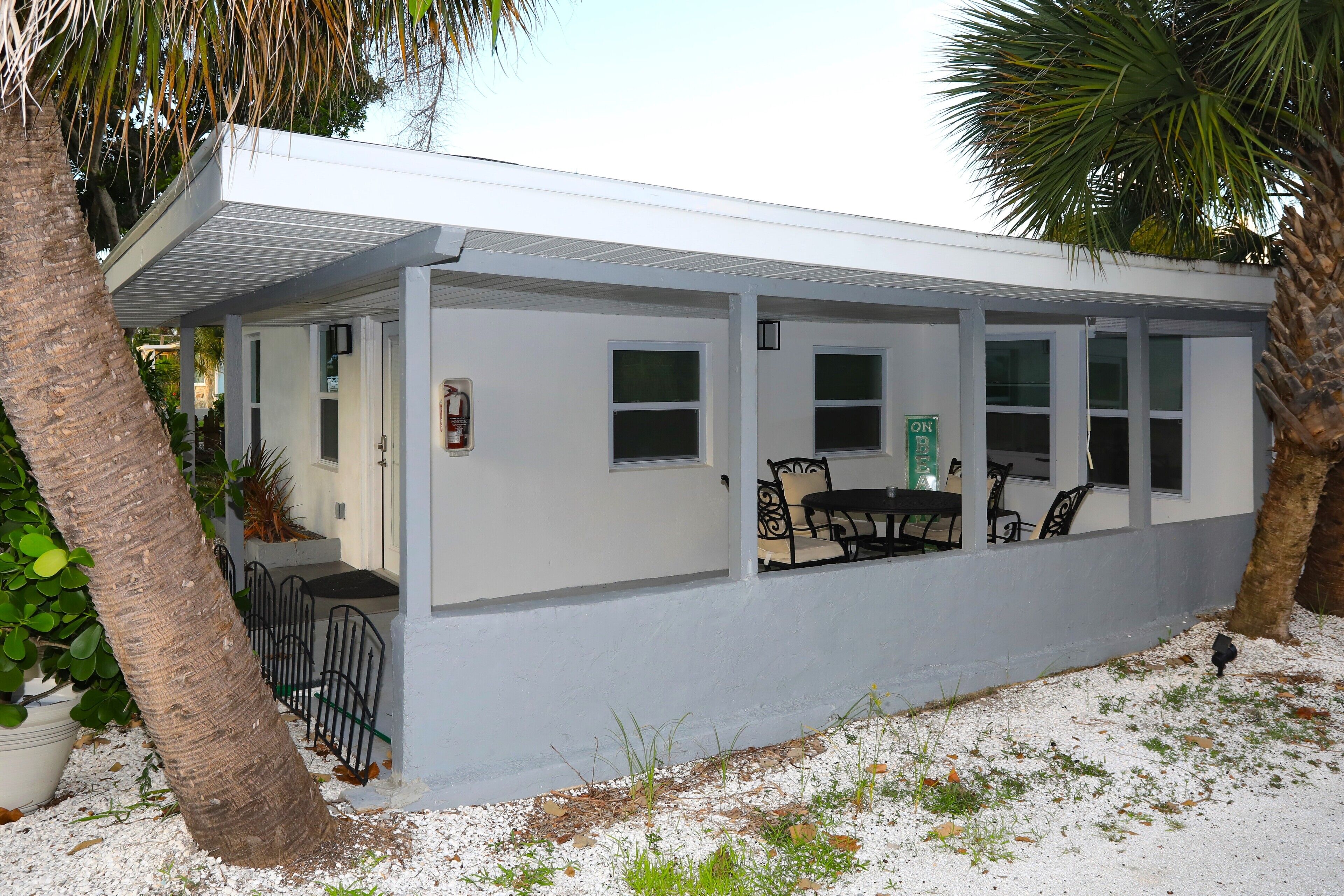 A covered outdoor seating area with a table and chairs, surrounded by white walls and palm trees.