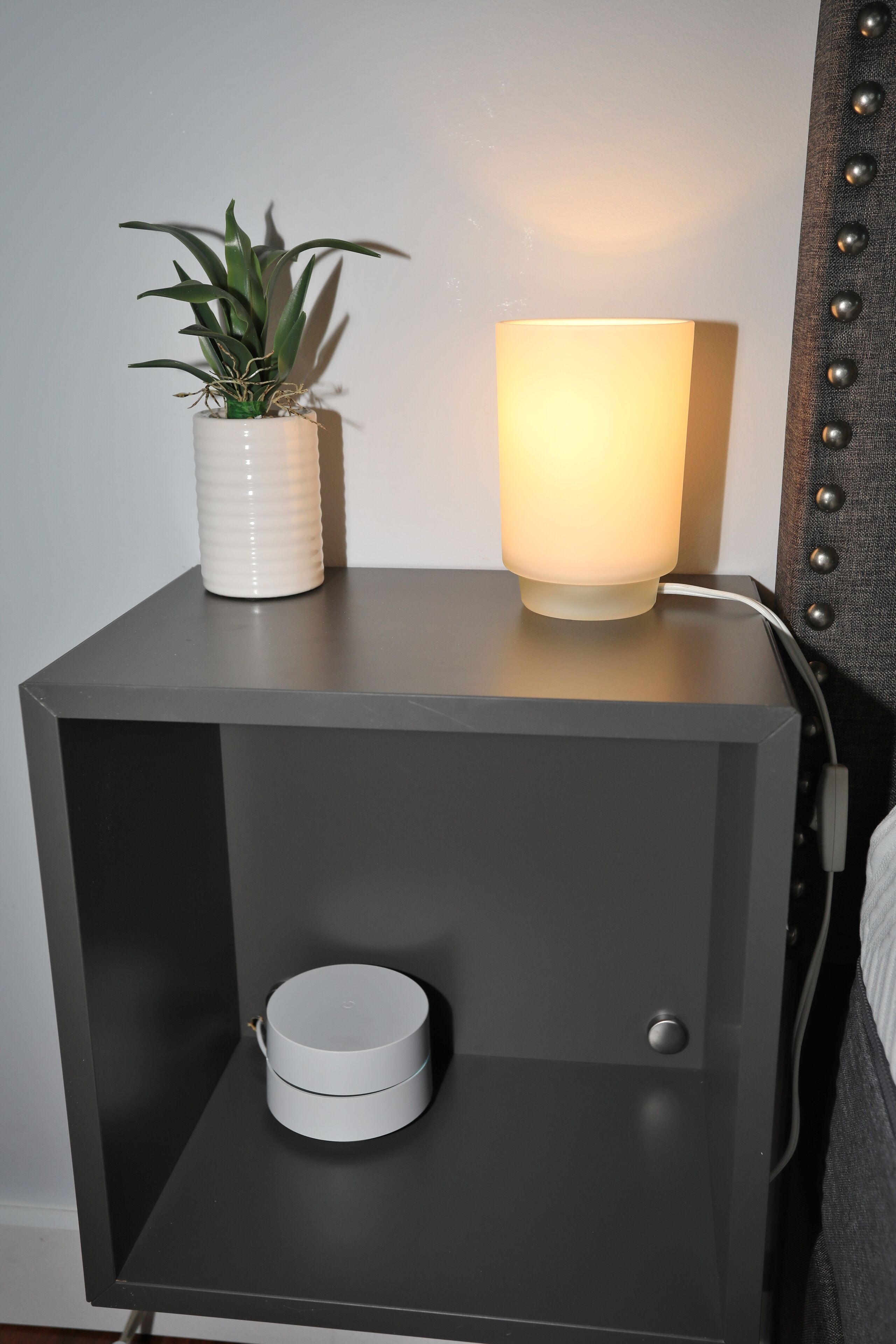 A modern bedside table with a white lamp, a small potted plant, and a white cylindrical object.