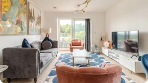 Un salon moderne avec un canapé gris, un fauteuil rose, une table basse et un téléviseur. Une grande fenêtre offre une vue sur la verdure à l’extérieur.