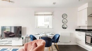 Une cuisine moderne avec des meubles blancs, un coin repas avec une table ronde et des chaises bleues, et un téléviseur dans l’espace salon.