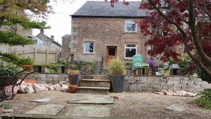 A stone house with a front door, steps leading to the entrance, and a garden area with plants and a wheelbarrow. - Cottage Hurst Green Sleeps 4, Views (Clitheroe)