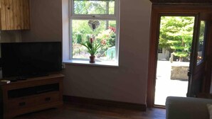 A living room with a television on a wooden stand, a window with a potted plant, and a view of a garden. - Cottage Hurst Green Sleeps 4, Views (Clitheroe)