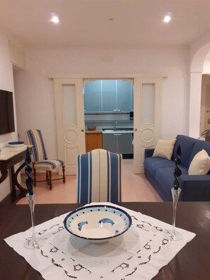 Uma sala de estar com uma mesa de jantar, cadeiras listradas, um sofá e uma cozinha ao fundo.