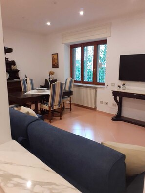 Uma sala de estar com um sofá azul, uma mesa de jantar com cadeiras, uma TV sobre um móvel e uma janela com vidro decorativo.