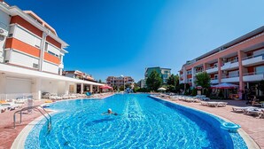 A swimming pool with a person swimming, surrounded by lounge chairs and umbrellas, with buildings in the background. - Studio - Sleeps 4 & Access to Pools (Bourgas)