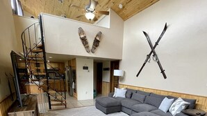 Un salon moderne avec un canapé d’angle gris, un grand fauteuil en forme de poire également gris et un plancher en bois. Un escalier avec une rampe en bois mène à l’étage supérieur.