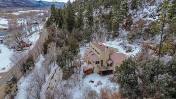 A large, multi-story house with a prominent balcony, surrounded by a snowy landscape and dense forest.