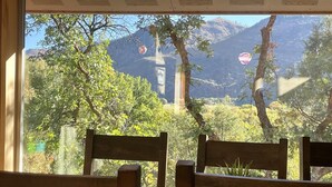 Une vue à travers une fenêtre, avec des chaises en bois au premier plan, une table recouverte d’un tapis tissé, et un paysage montagneux avec des arbres et une montgolfière en arrière-plan.