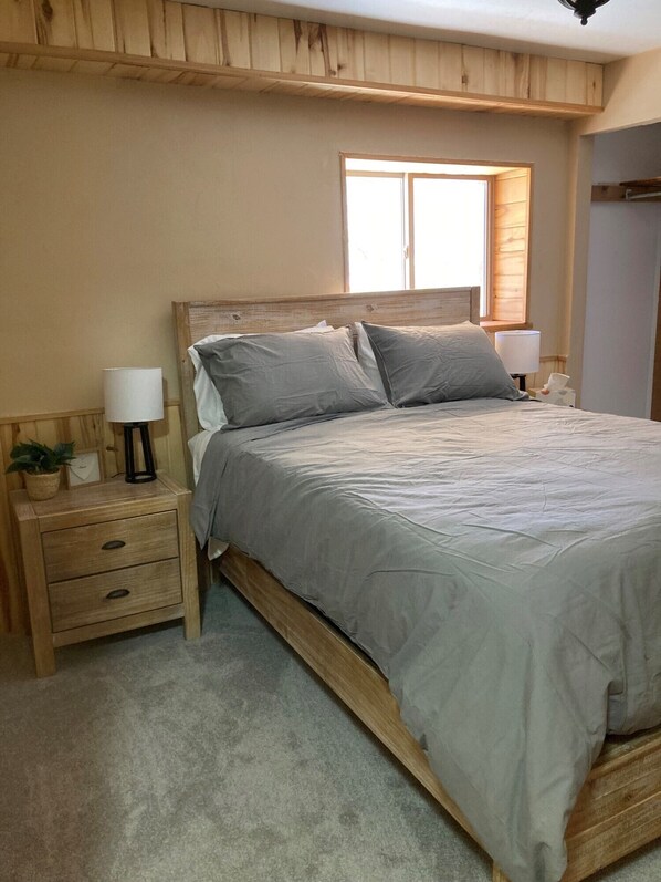 Un dormitorio con estructura de cama de madera, dos mesitas de noche, una ventana y una planta pequeña.
