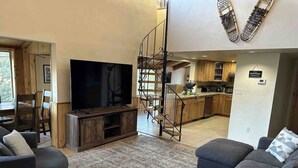 Un salon doté d’un grand téléviseur à écran plat, de canapés gris et d’un escalier menant à la cuisine.