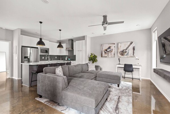 Un salon moderne avec un canapé d’angle gris, une cuisine avec des meubles blancs, un coin repas avec une table blanche et des œuvres d’art accrochées aux murs.