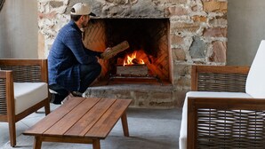 Ein Kamin aus Stein mit darüber angebrachten Geweihen, eine Person sitzt auf einer Holzbank, und Korbmöbel in einem Raum mit Betonboden.