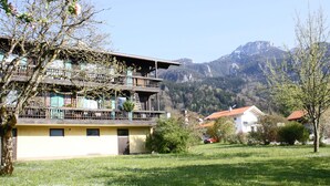 Un edificio a più piani con balconi, immerso nel verde tra prati e alberi, con montagne sullo sfondo.