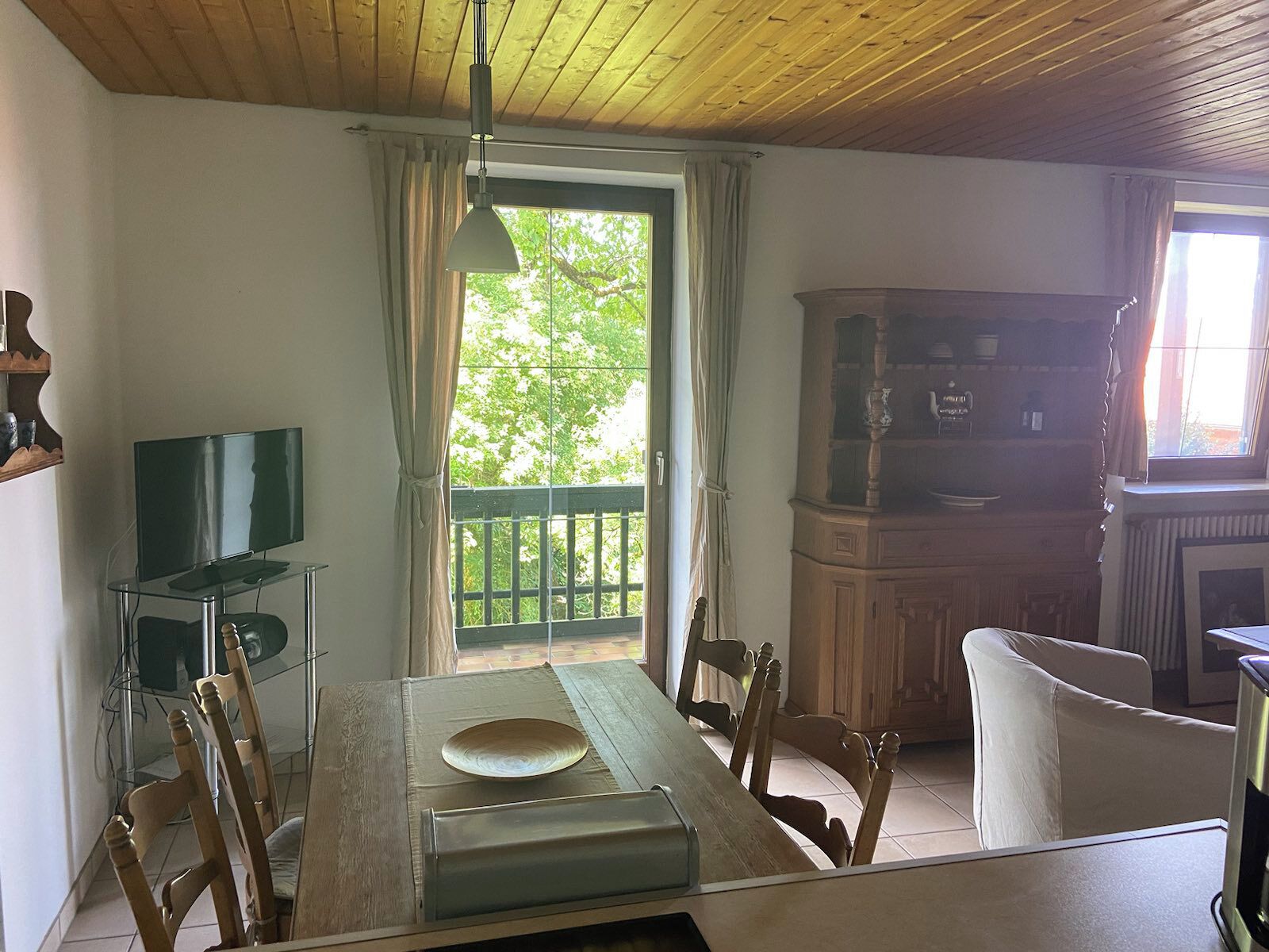 Un área de comedor con una mesa de madera, sillas, un televisor sobre un pedestal de vidrio y vistas a la vegetación por una ventana.