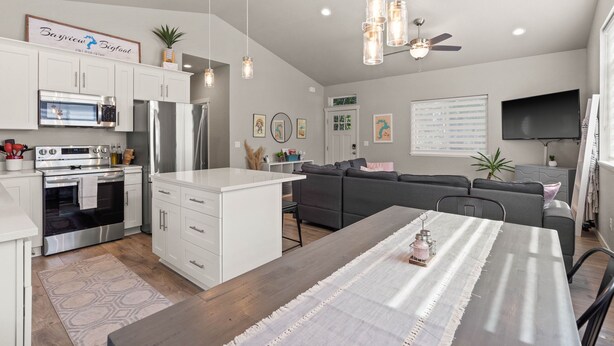 A modern kitchen with white cabinets, a central island, and stainless steel appliances. There is a dining area with a table and chairs, a gray sofa, and a ceiling fan. - PNW ~ Bayview Bigfoot steps to Lake Pend Oreille/Hot Tub (Bayview)