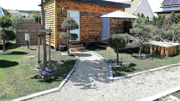 Eine kleine Holzhütte mit einem Kiesweg, der zum Eingang führt, umgeben von einem gepflegten Garten mit verschiedenen Sträuchern und einem kleinen Baum.