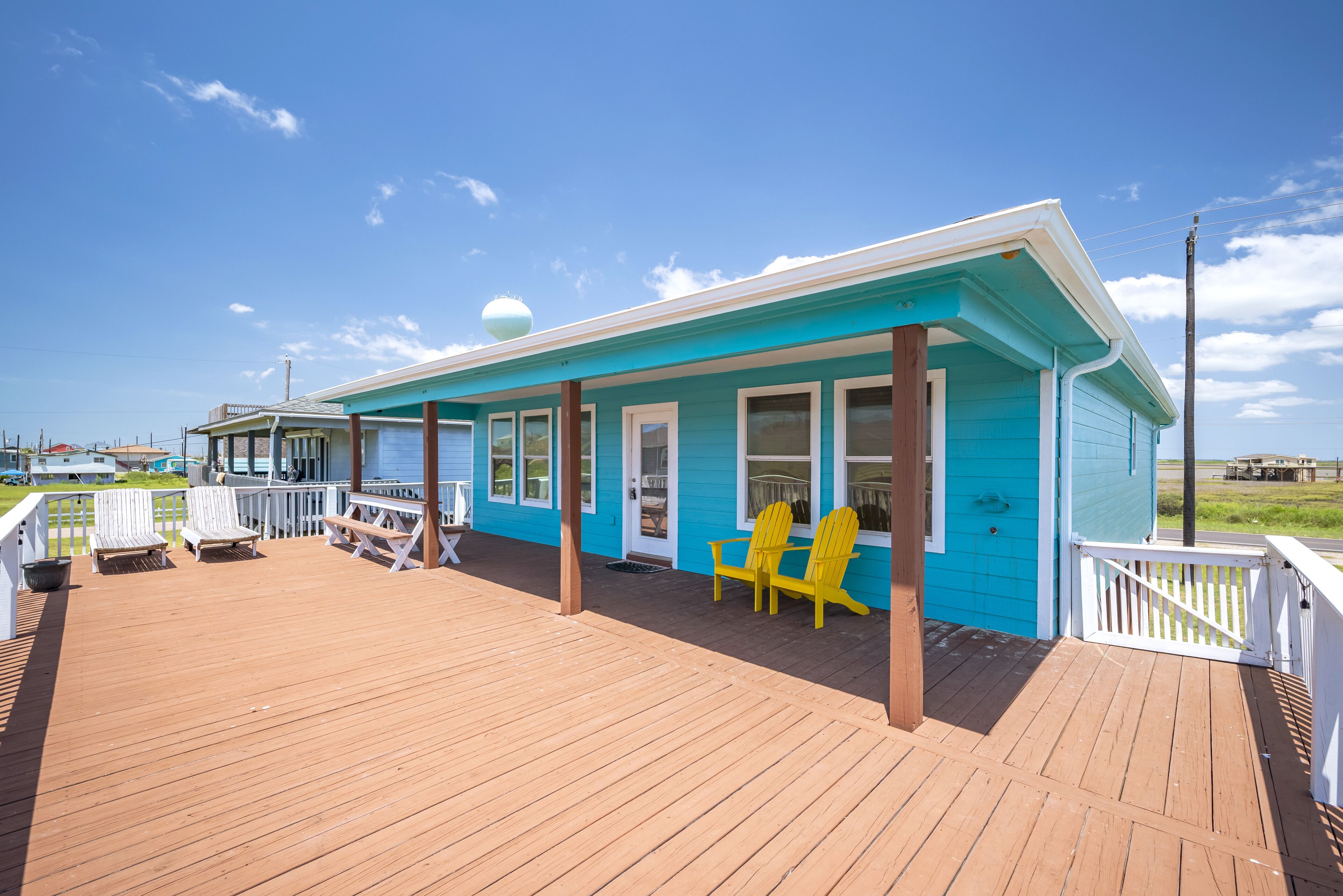 A blue building with a wooden deck, white railings, and yellow Adirondack chairs.