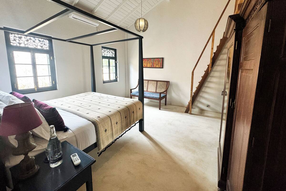 A bedroom with a four-poster bed, a desk with a lamp, a chair, and a staircase.