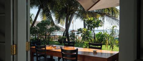 An outdoor dining area with a wooden table and chairs, surrounded by lush greenery and a view of the ocean.