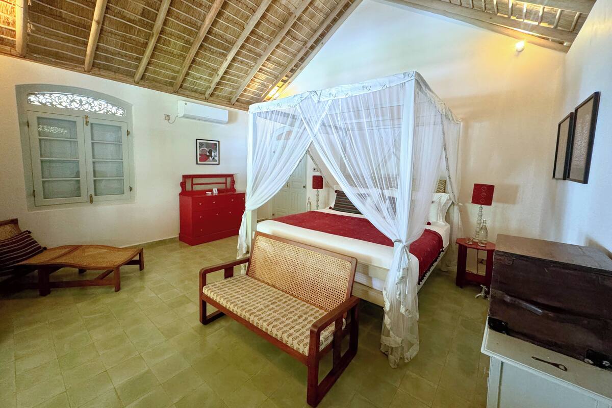 A bedroom with a four-poster bed, a wooden bench, a red dresser, and a small table with a lamp.