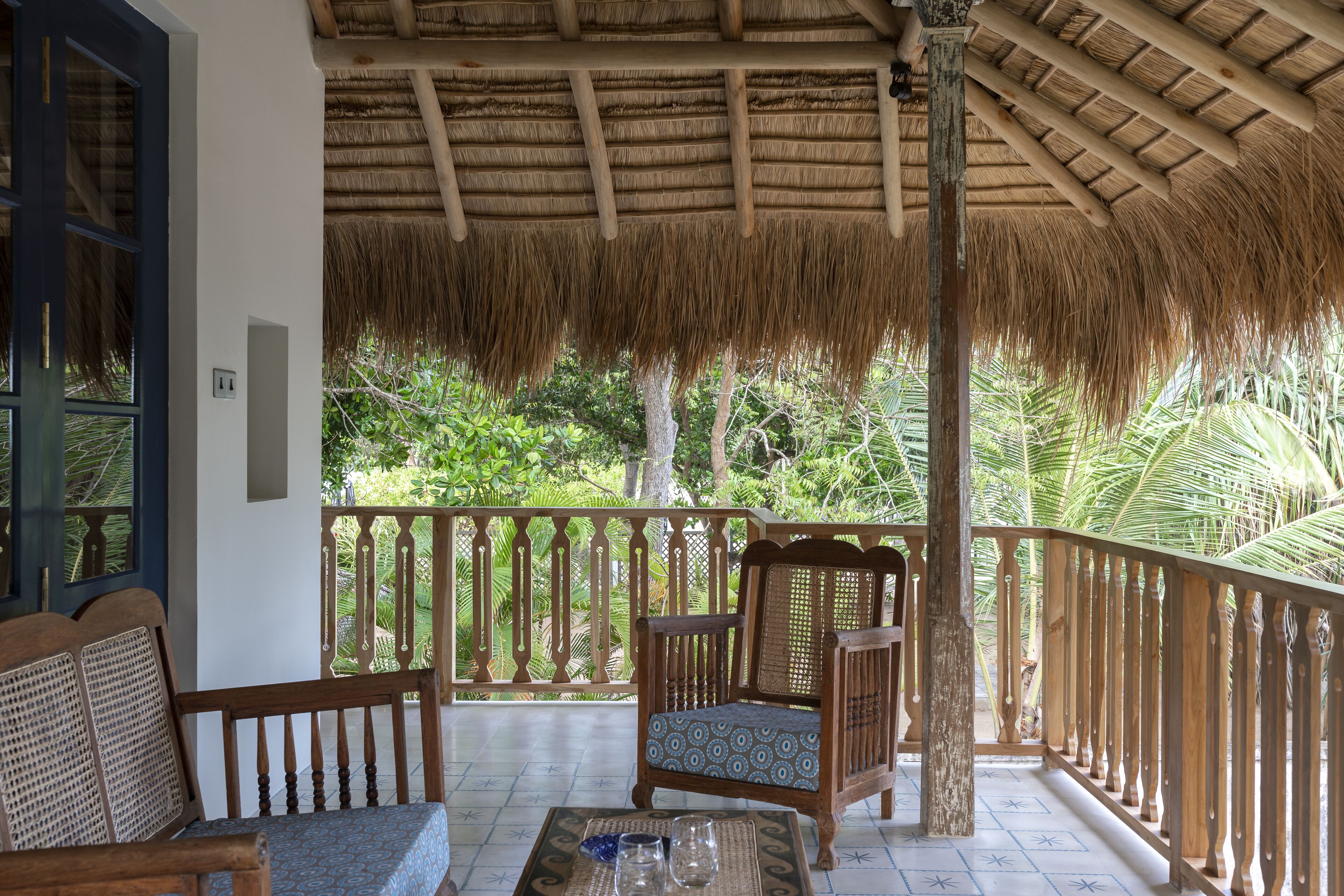 A covered porch with a thatched roof, wooden furniture, and a view of lush greenery.