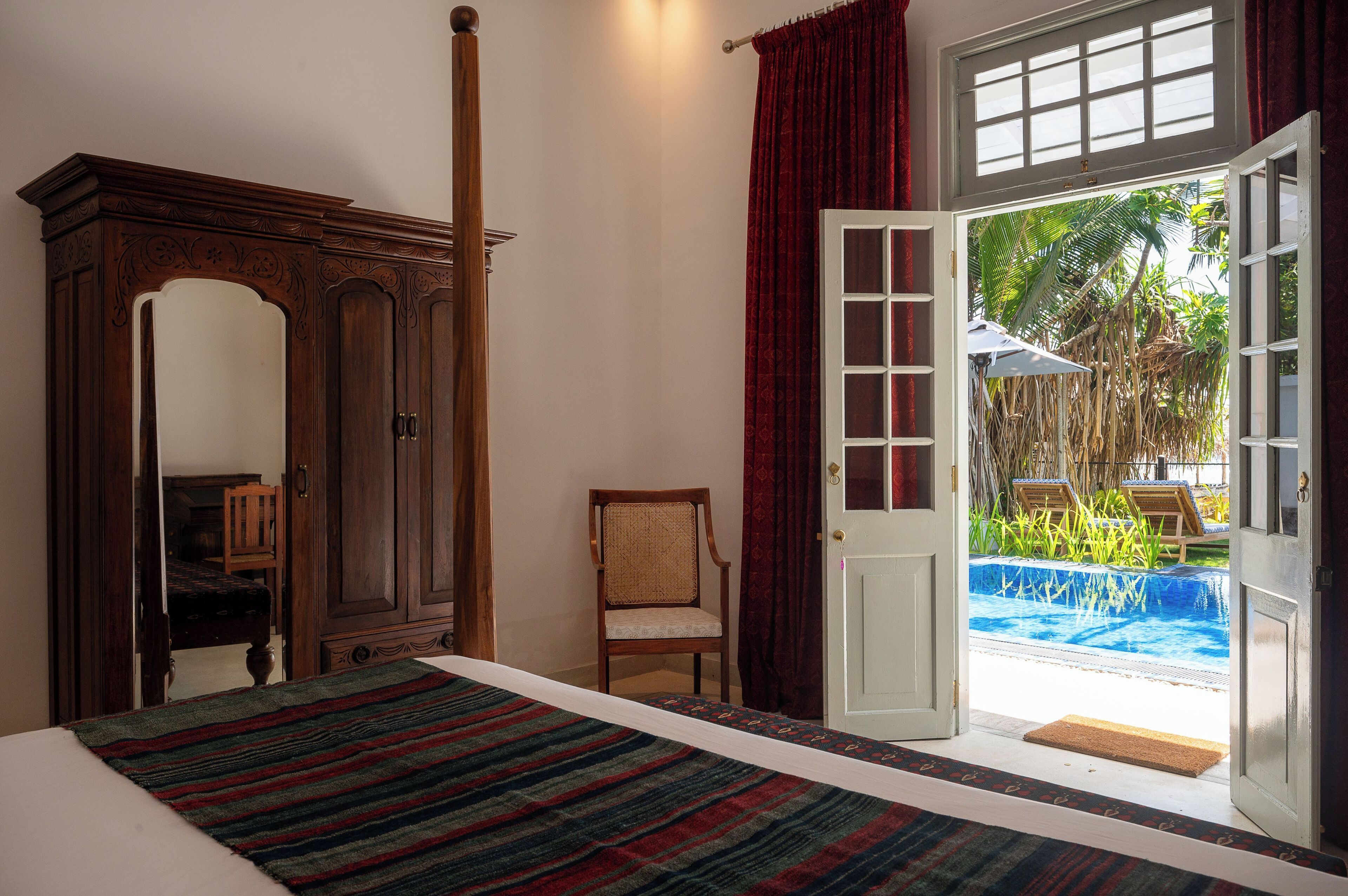 A bedroom with a bed, a wooden wardrobe, a chair, and a door leading to a pool area.