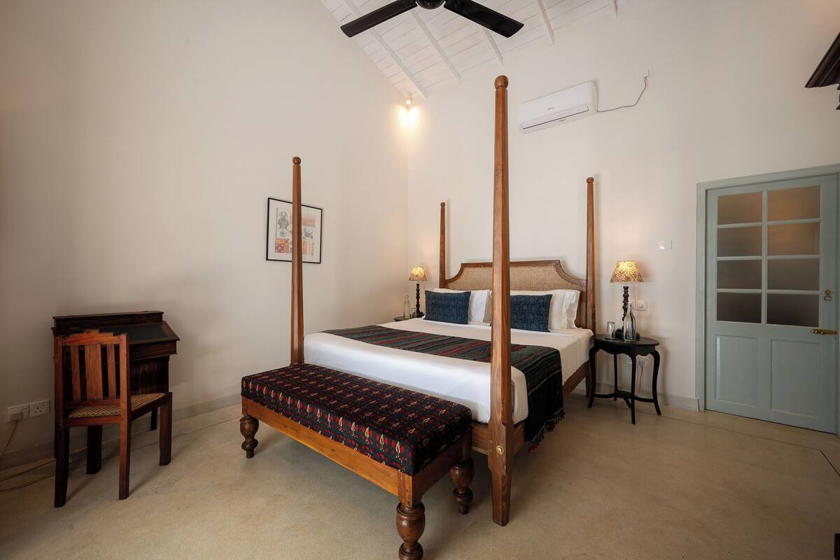 A four-poster bed with a bench, a desk, and a chair in a room with a door and an air conditioning unit.