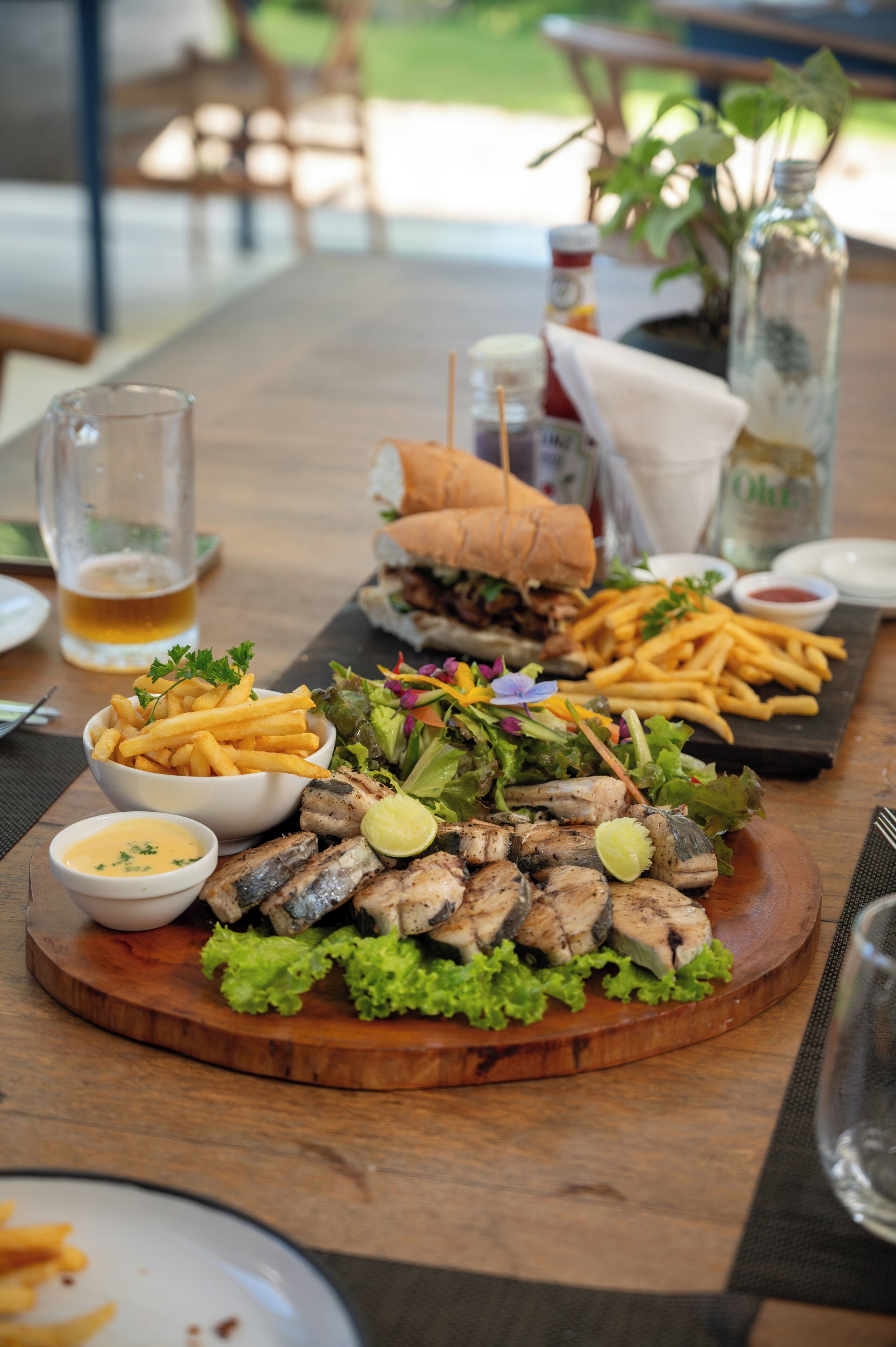 A wooden table with a variety of food including grilled fish, fries, a salad, and a sandwich. There are also condiments and a bottle of water on the table.