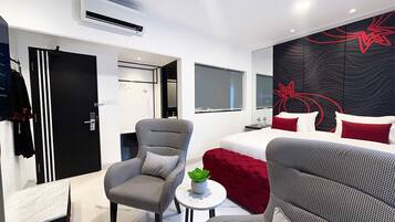 Una habitación de hotel moderna con una cama, un sillón gris, una mesita redonda con una planta y un sofá rojo.