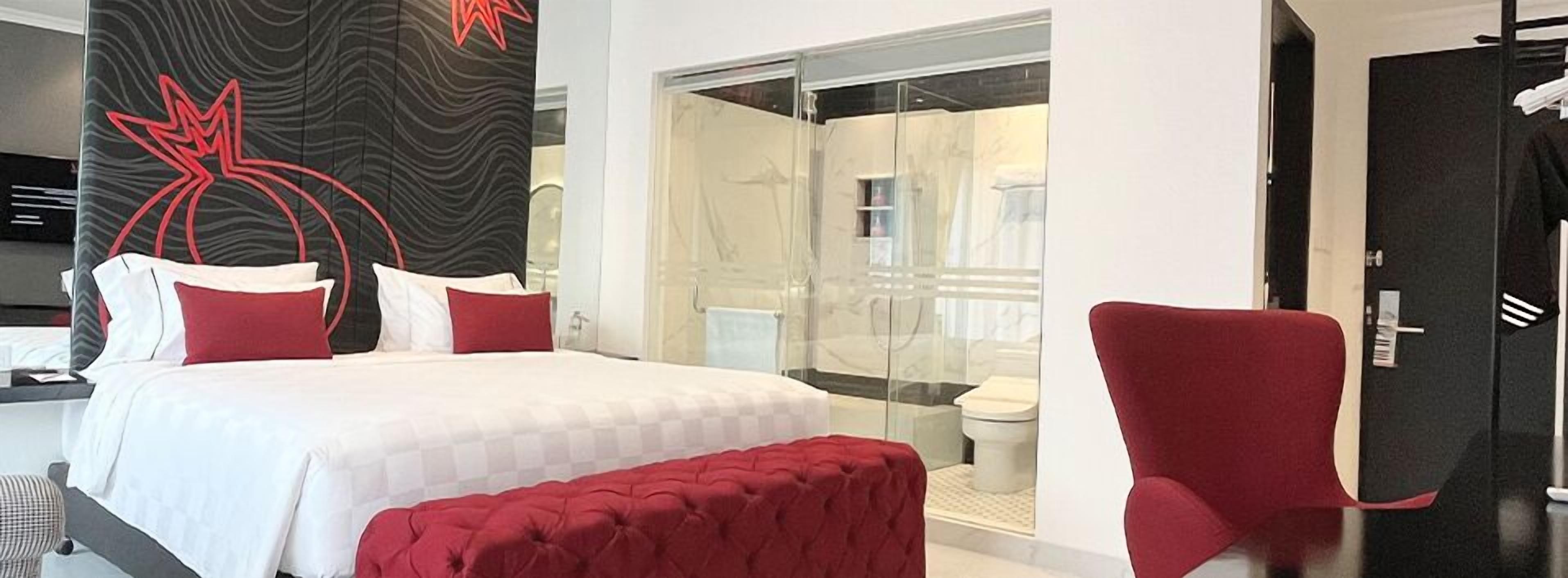 Een moderne hotelkamer met een groot bed, rode en witte kussens, een rode poef, een douchecabine van glas en een zwarte deur.