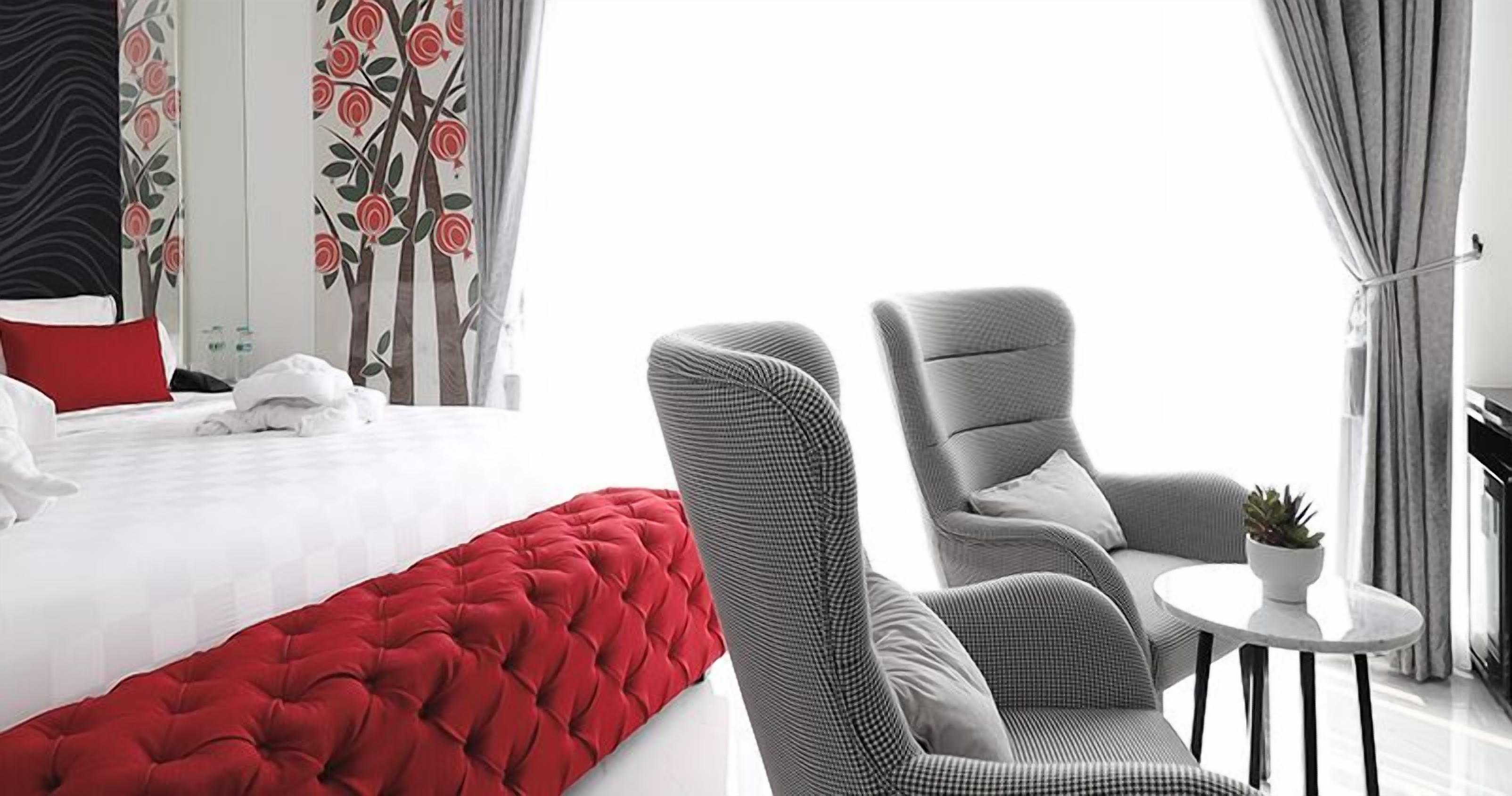 Hotelkamer met een groot bed, een rode decoratiekussen, een grijze fauteuil, een klein rond tafeltje met een plant en gordijnen met een bloemenprint.