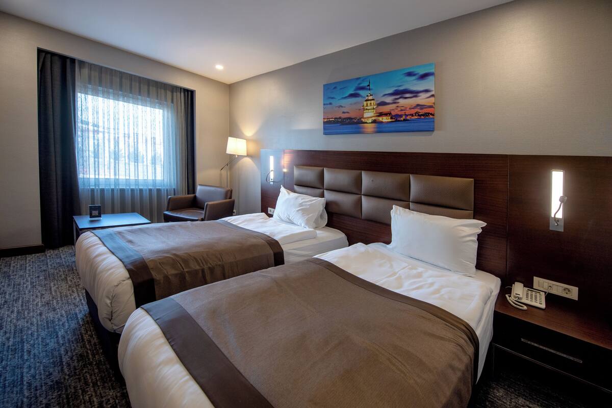 A hotel room with two beds, a desk, a chair, a lamp, a painting, and a phone on the nightstand.