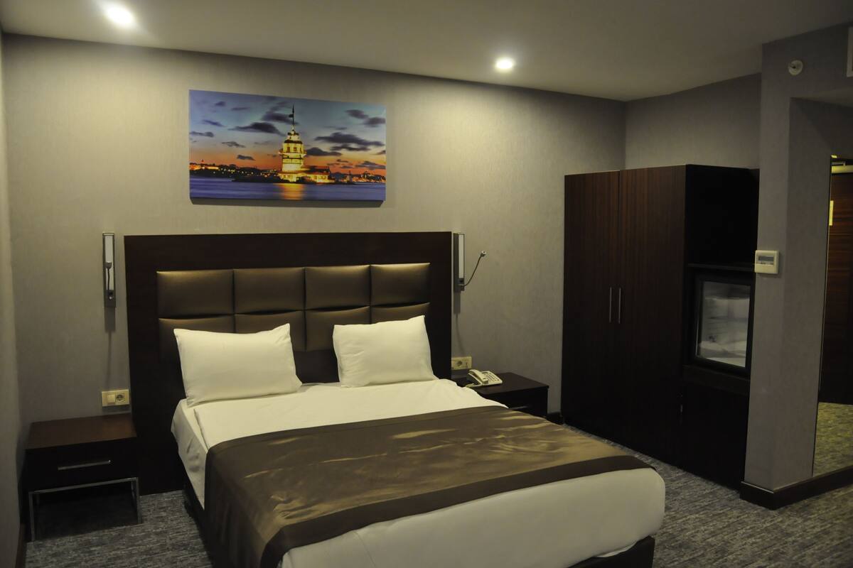 A modern hotel room with a large bed, a nightstand, a wardrobe, and a painting of a cityscape on the wall.