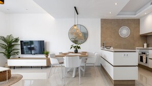 Une cuisine moderne avec des meubles blancs, un coin repas comprenant une table en bois et des chaises blanches, un grand téléviseur à écran plat et un élément décoratif mural.