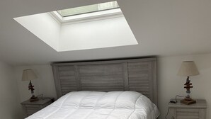 Un dormitori amb una finestra panoràmica, un llit amb edredó blanc, dues tauletes de nit amb làmpades i un capçal de llit de fusta.