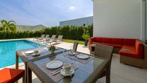 A dining area with a pool view, outdoor furniture, and a modern building in the background.