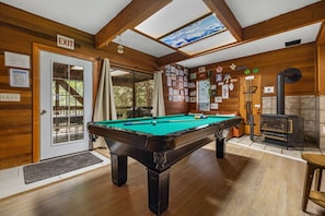 A pool table in a room with wooden walls, an exit sign, a skylight, and a fireplace.