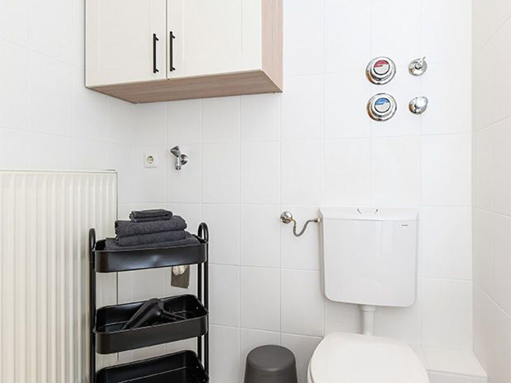A compact bathroom with a white toilet, a black shelf unit with towels, a radiator, and a wall-mounted toilet paper holder.