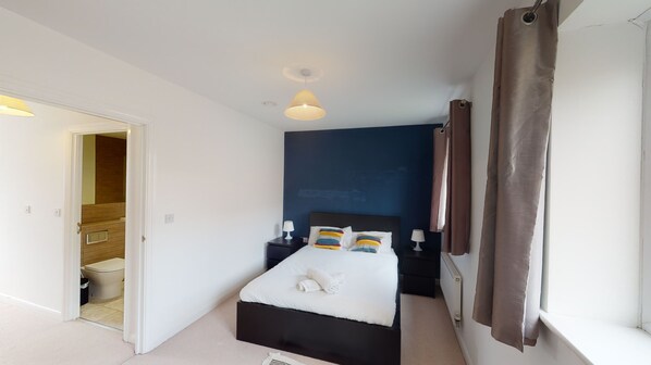 A bedroom with a bed, bedside tables, a nightstand, a window with curtains, and a door leading to a bathroom. - The Central Lodge II (Dartford)