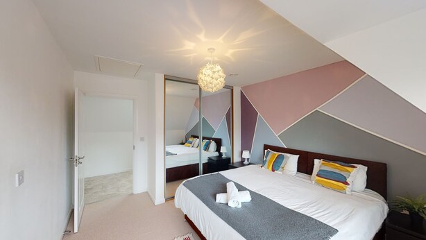 A modern bedroom with a large bed, a bedside table, a nightstand, a mirror, and a unique ceiling light fixture. - The Central Lodge II (Dartford)
