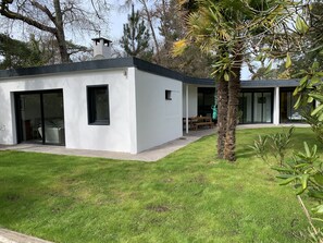 Een modern huis met twee verdiepingen, een plat dak, grote glazen schuifdeuren en een klein terras met een tafel en stoelen.