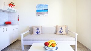 A cozy living area with a white sofa, nautical-themed cushions, a small table with a fruit bowl, and a kitchen with white cabinets and a red appliance. - Beachfront Apartments in Gouves (Hersonissos)