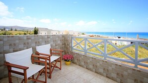 A balcony with two wooden chairs and a table, a yellow flower pot, and a view of the sea and distant buildings. - Beachfront Apartments in Gouves (Hersonissos)