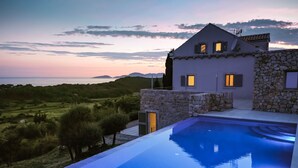 A modern villa with a swimming pool, stone walls, and a view of the sea at sunset.