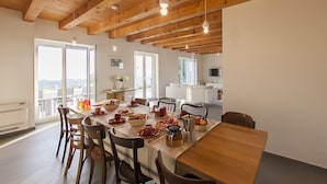 A dining area with a wooden table set for a meal, surrounded by chairs. There are large windows providing natural light and a view of the outdoors.