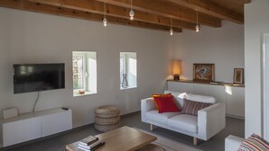 A modern living room with a white sofa, a wooden coffee table, and a flat-screen TV mounted on a white cabinet. There are woven baskets on the floor, a lamp on a side table, and a painting on the wall.