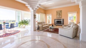 Un salon spacieux avec un canapé beige, un fauteuil blanc et une cheminée. On y trouve également un coin repas avec une table et des chaises, ainsi qu’un balcon avec vue.
