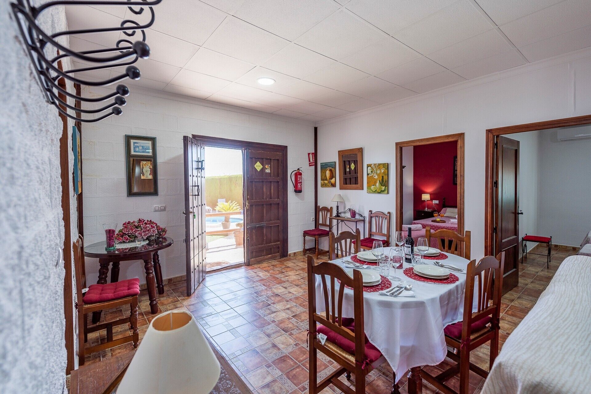 Un coin repas avec une table ronde dressée pour quatre personnes, des chaises en bois, une nappe blanche et un coussin rouge sur l’une des chaises.