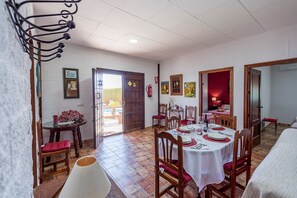 Un comedor con una mesa redonda preparada para cuatro personas, sillas de madera, mantel blanco y un cojín rojo en una de las sillas.
